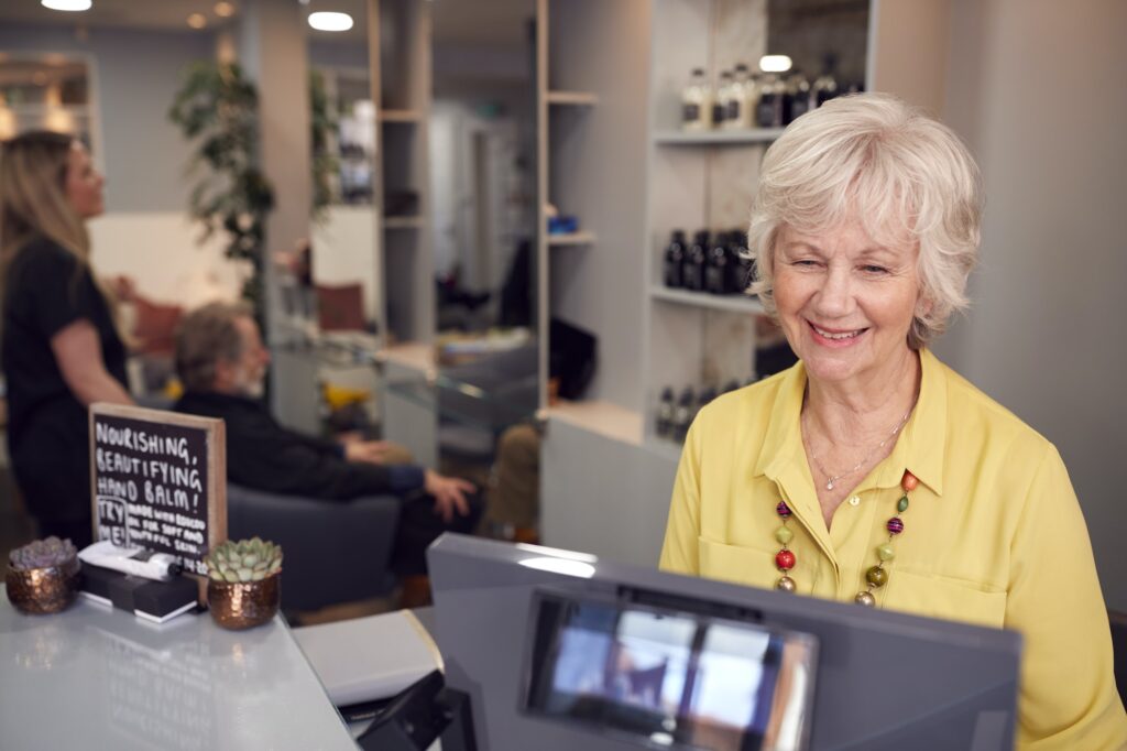 Senior Woman Arriving For Appointment To Have Hair Cut In Hairdressing Salon At Reception Desk