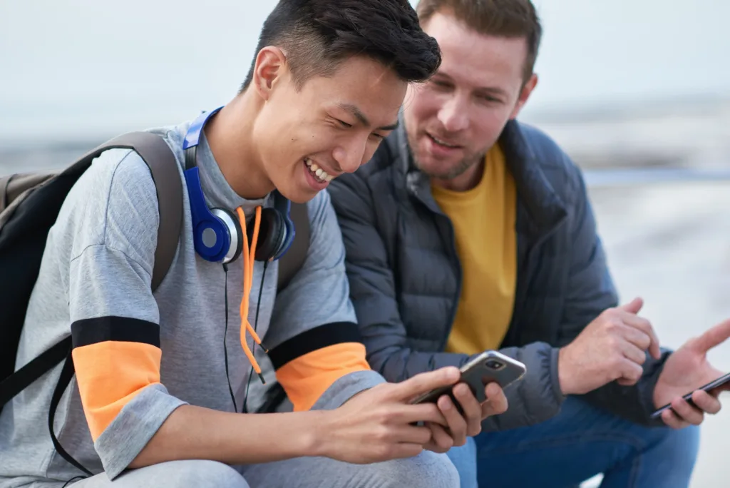 Friends using smartphones hanging out by seaside browsing social media messages texting with mobile