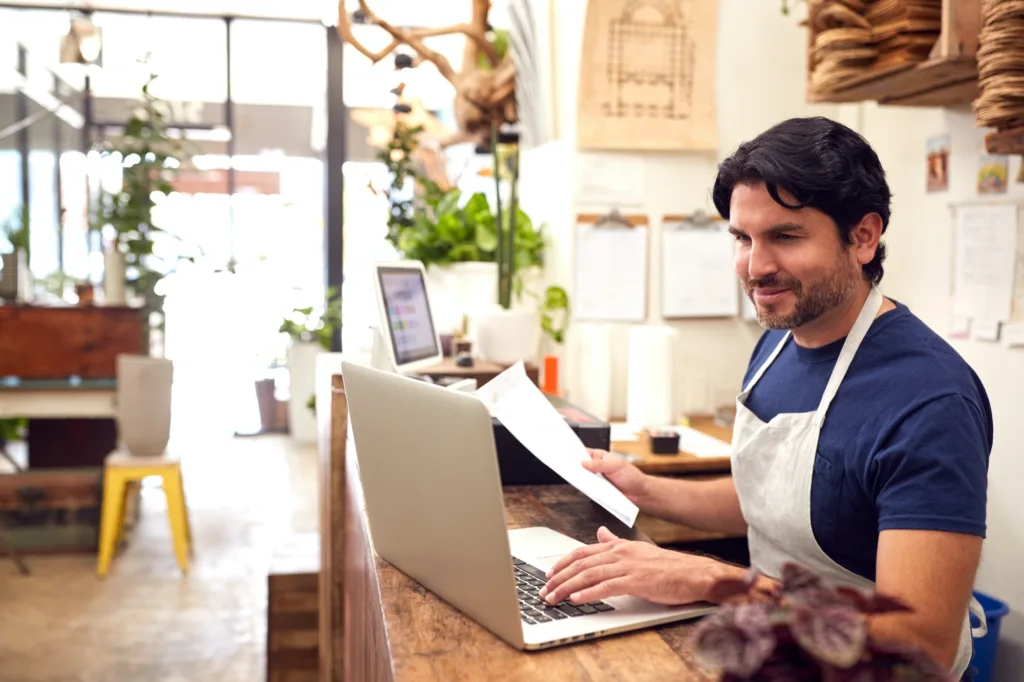 Male Sales Assistant Working On Landing Page Behind Sales Desk Of Florists Store